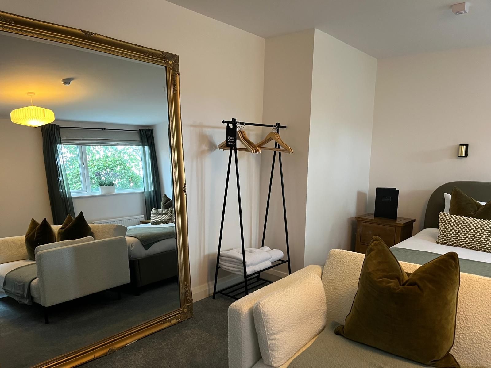 Modern bedroom with beige bed, large gold mirror, and lounge chair reflected in the mirror