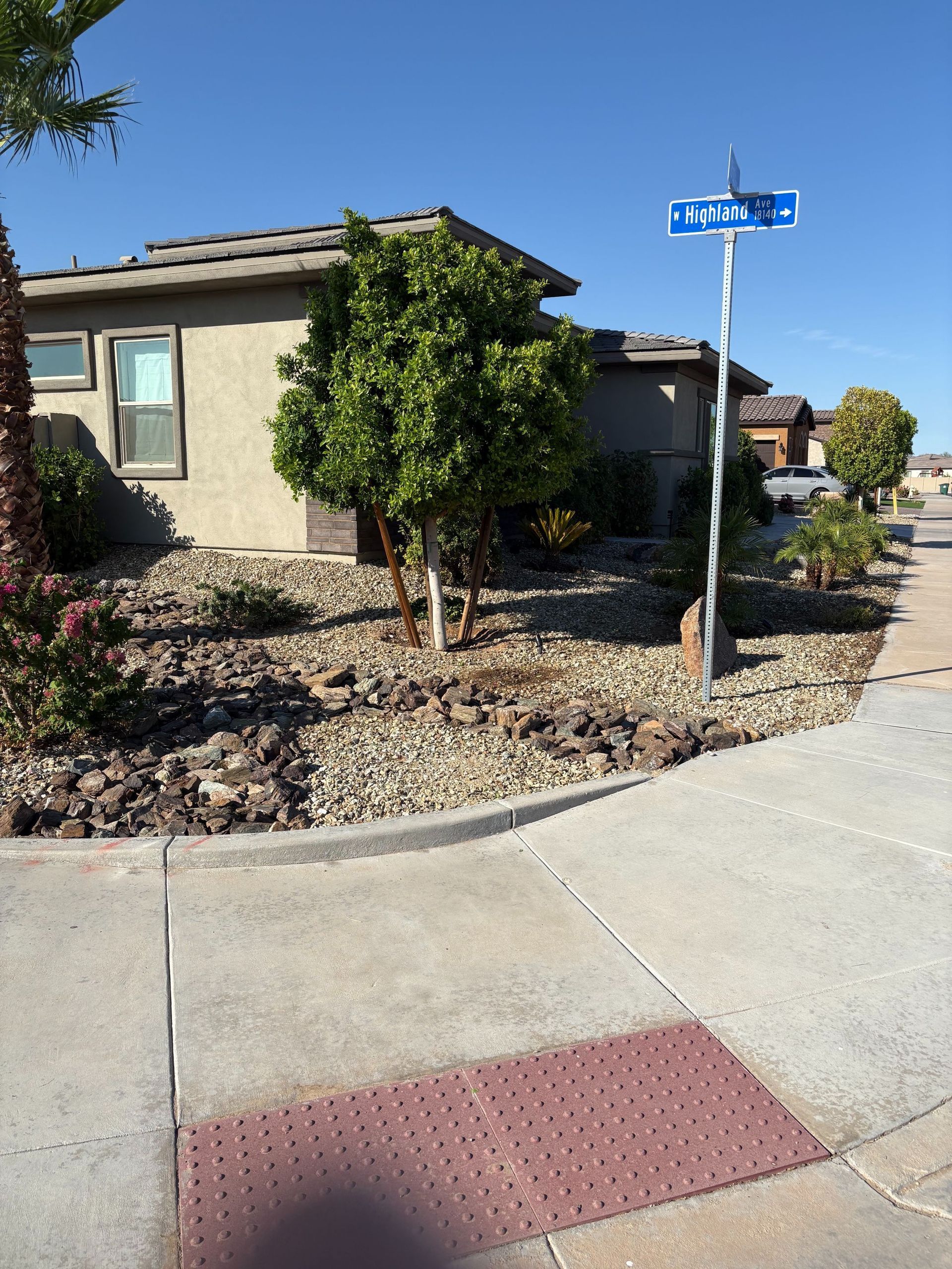 A house at a corner with a sidewalk. Street sign. Stone landscaping and green shrubbery. Blue sky.