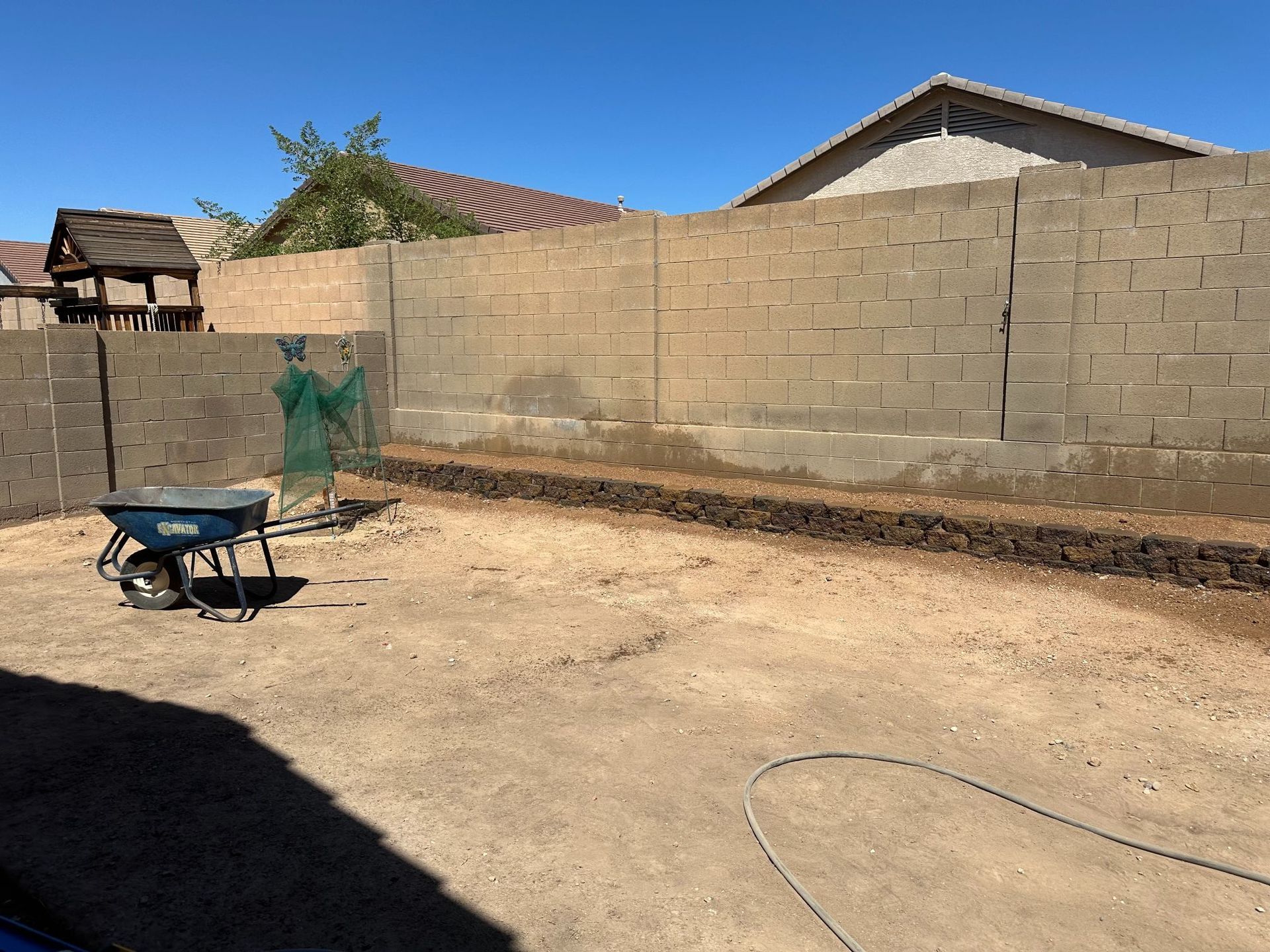 A sunny backyard with a wheelbarrow, brick wall, and a dug trench.