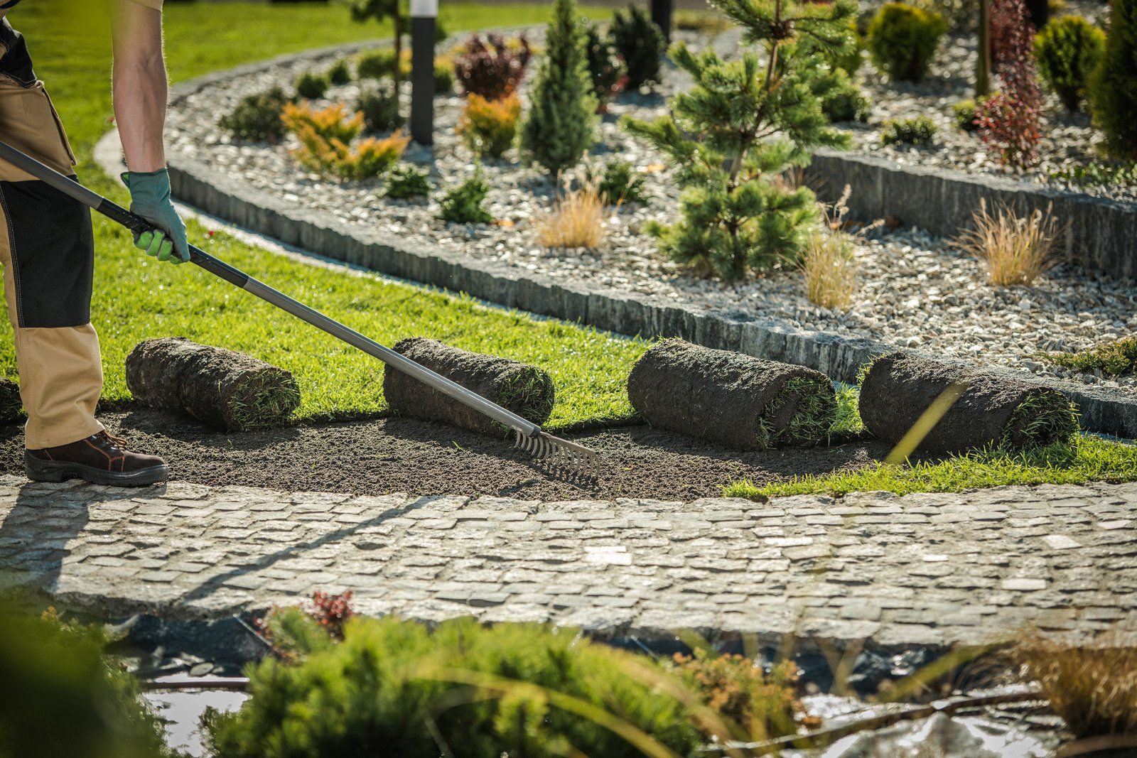 A person laying sod rolls with a rake in a garden bed. Green and gray landscaping features are visible.