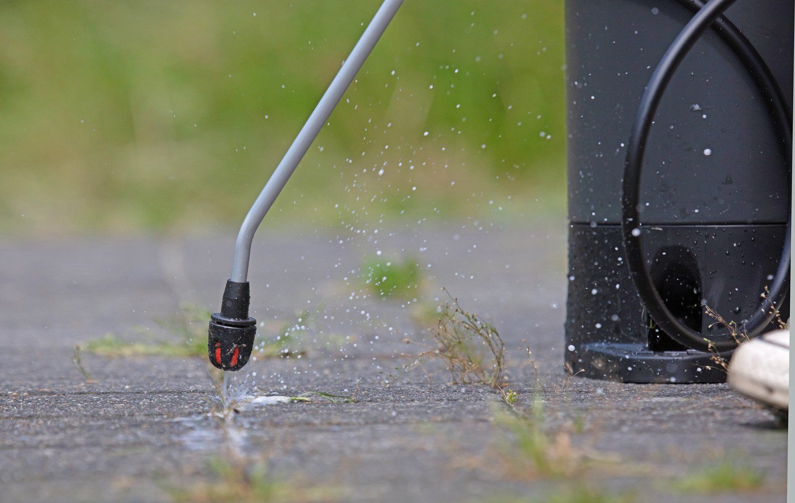 A spray nozzle dispensing liquid on a concrete surface; droplets are visible. A black container and hose are also present.