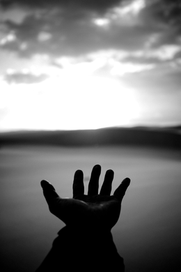 Silhouette of a hand reaching upward towards a bright, cloudy sky over a body of water.