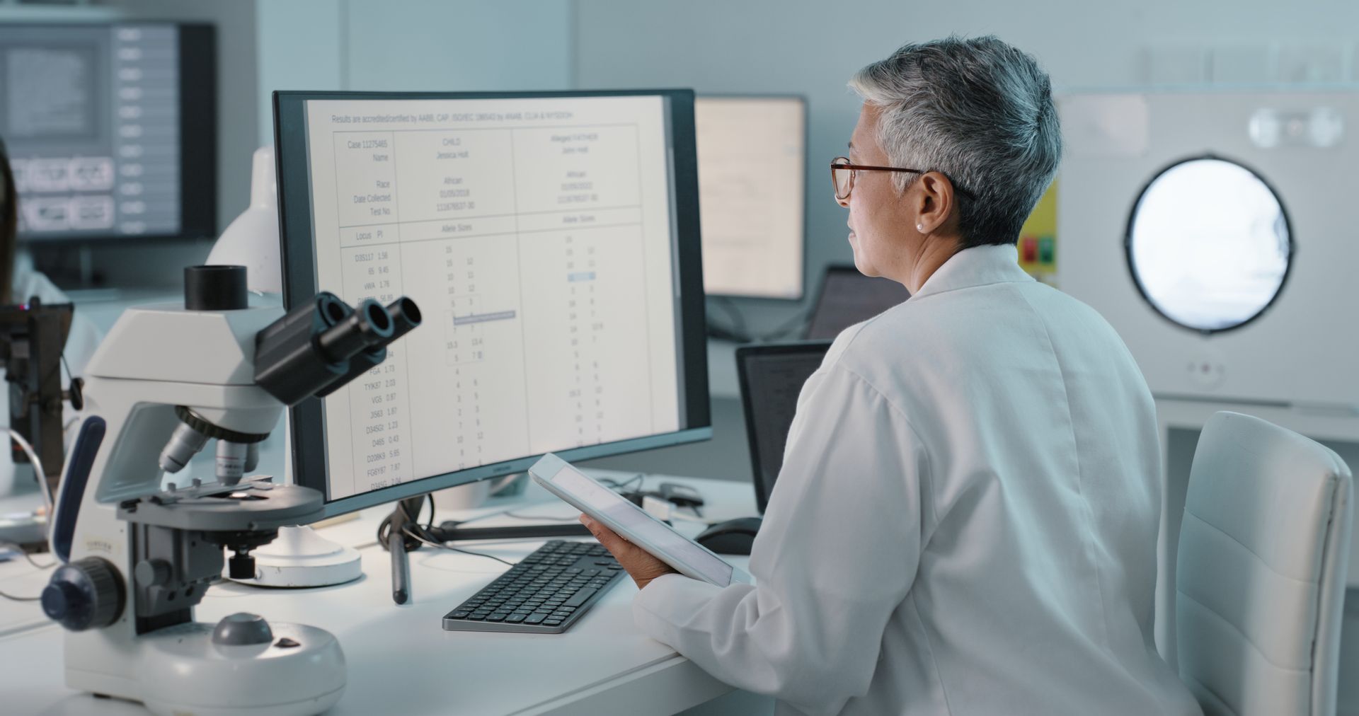 A scientist is sitting at a desk in front of a computer and a microscope.