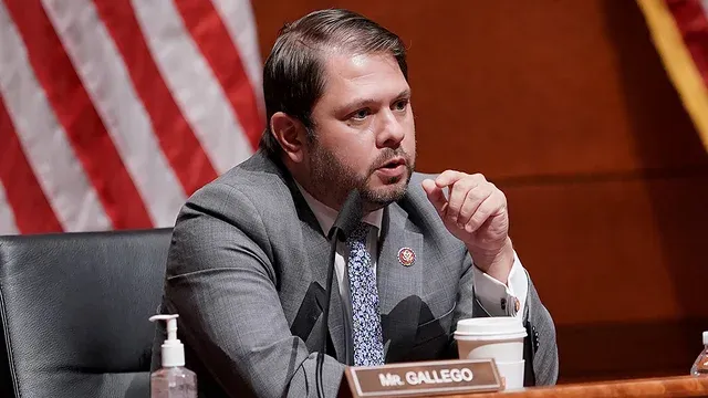 A man in a suit and tie is sitting at a table in front of an american flag.