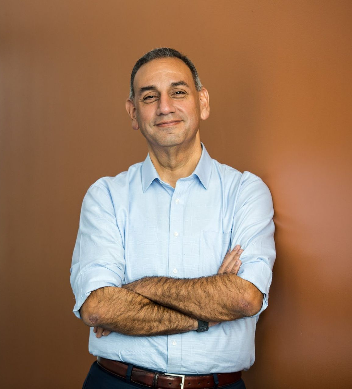 Man standing with arms crossed, wearing a light blue shirt against a brown background