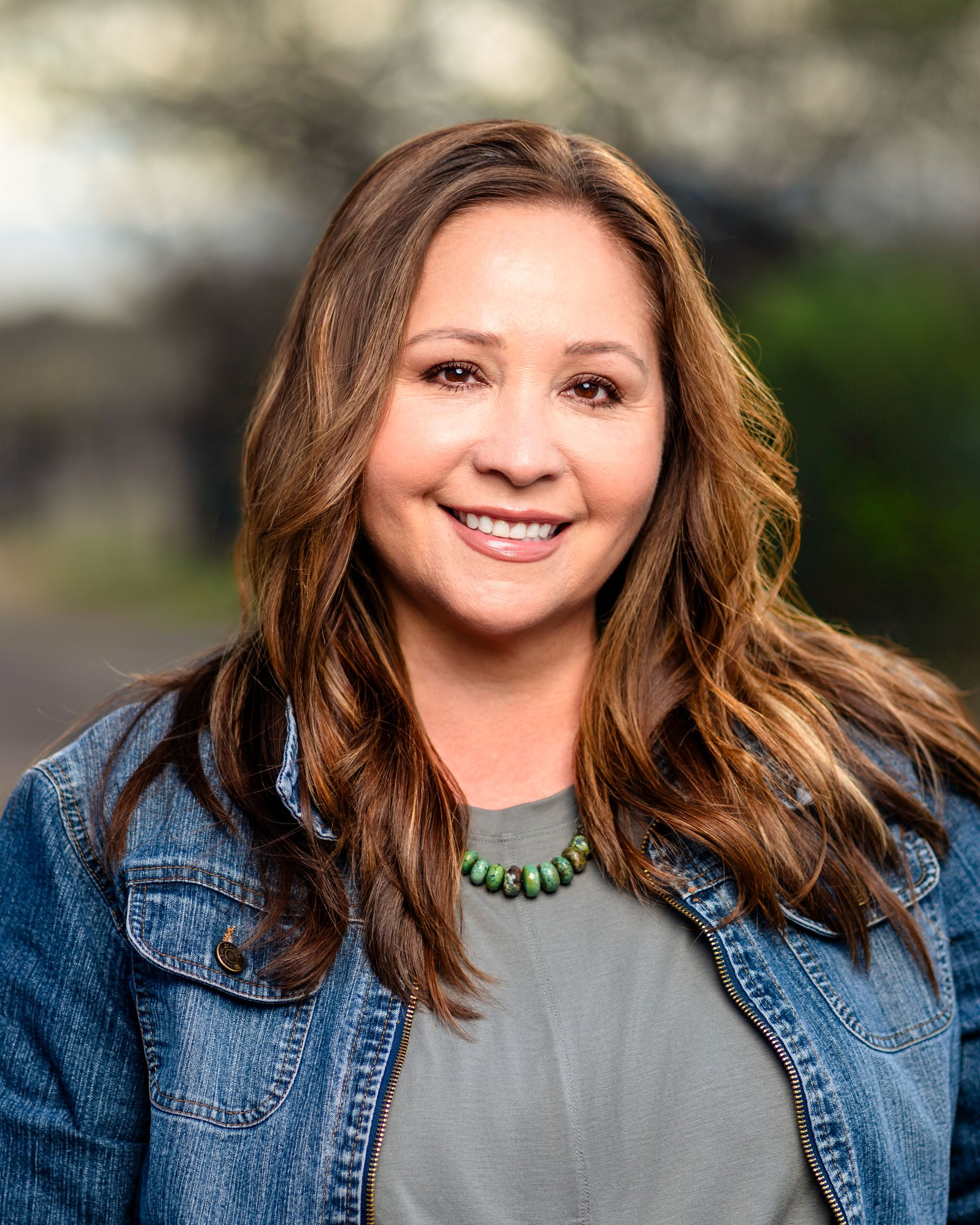 Smiling woman in a denim jacket and gray top, standing outdoors with a blurred background.