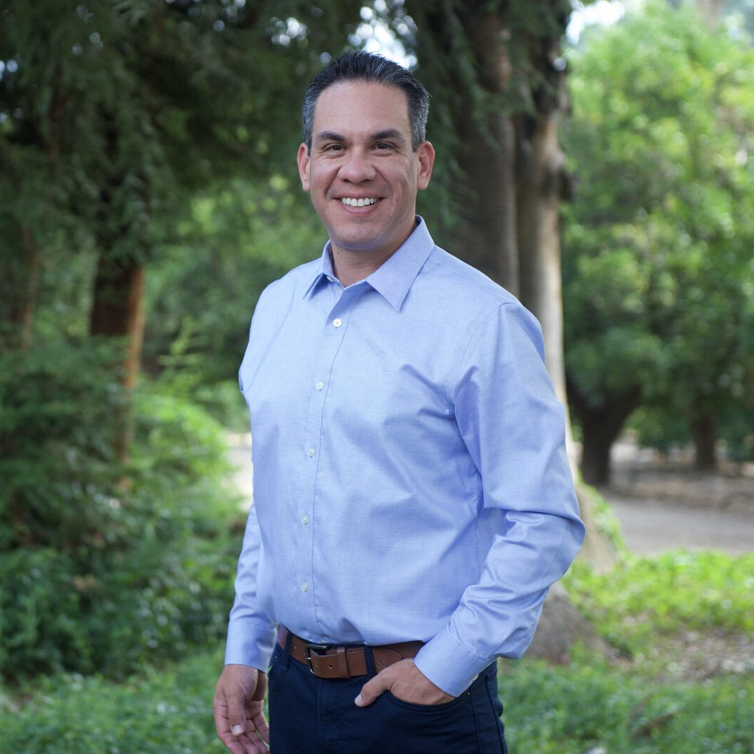 A man in a blue shirt is standing in a park with his hands in his pockets