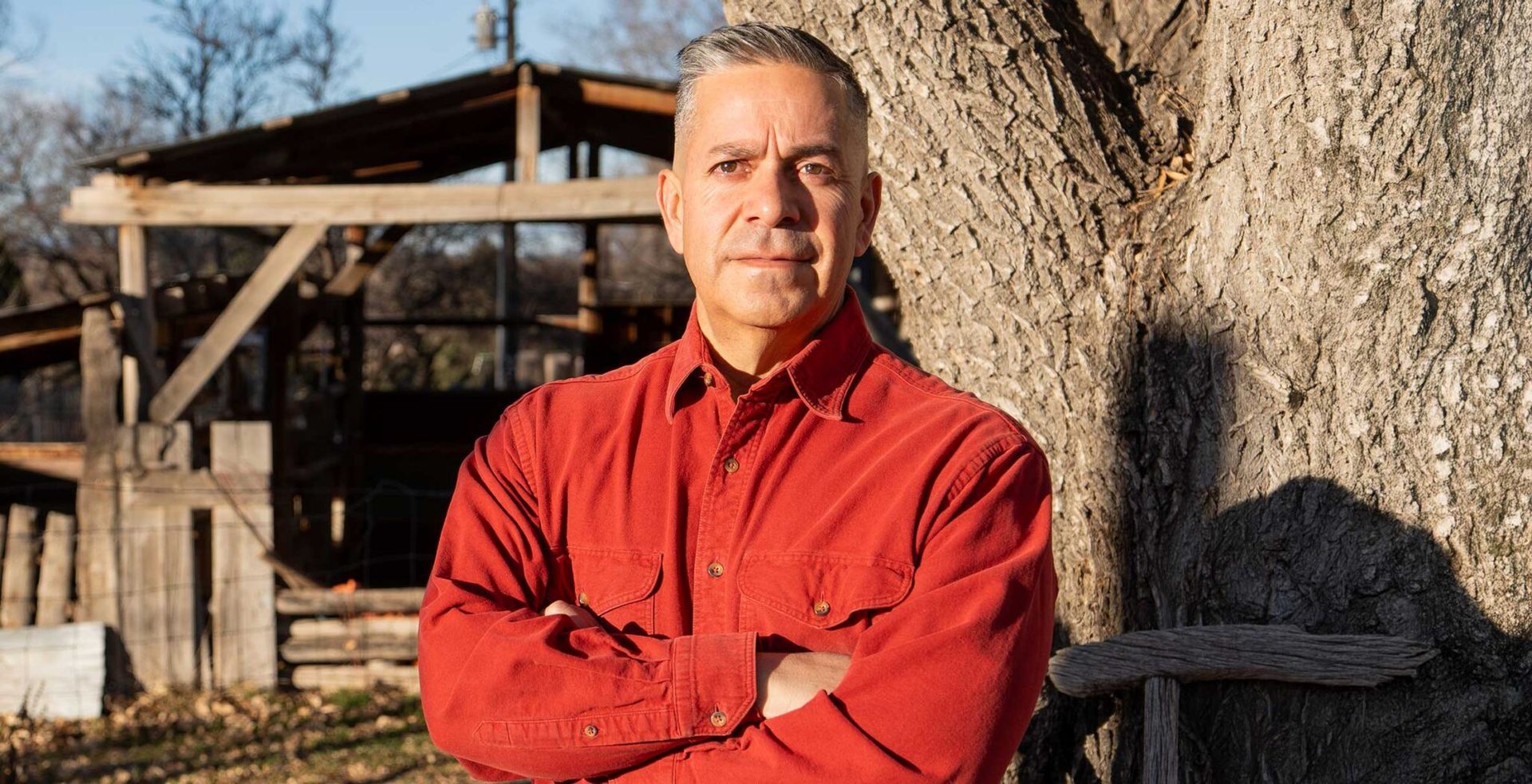 A man in a white shirt stands with his arms crossed in front of a tree
