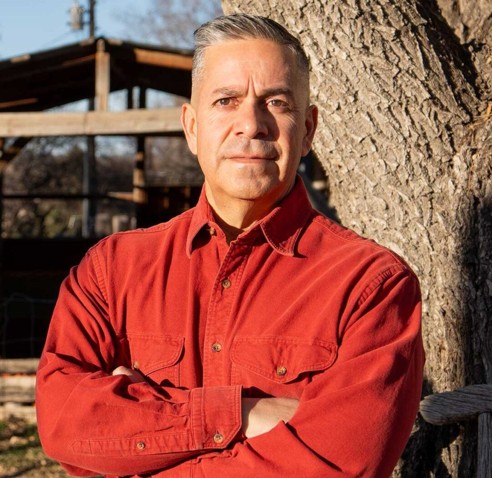 A man in a white shirt stands with his arms crossed in front of a tree