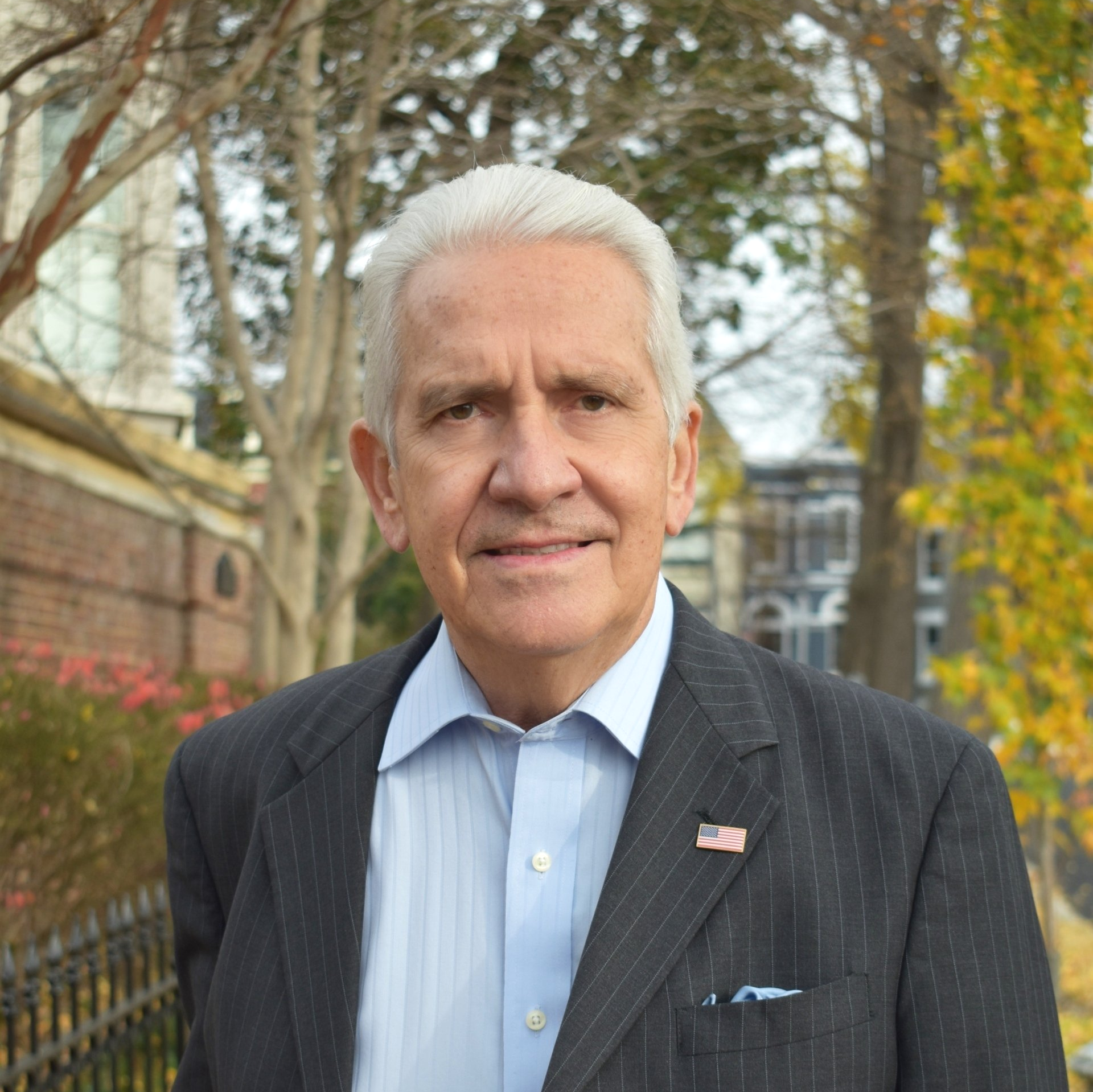 A man in a suit and blue shirt is standing in front of a fence.