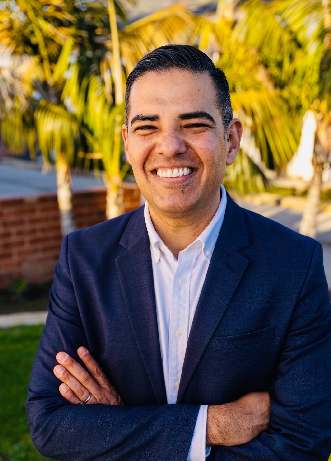 A man in a suit and shirt is standing in front of a tree.