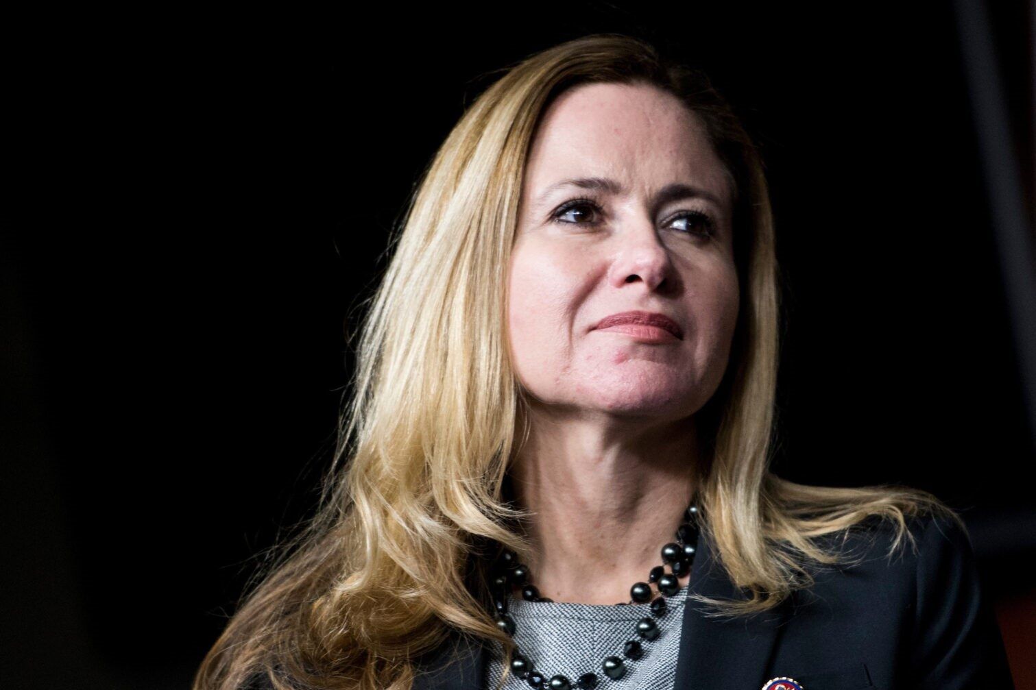 A woman with long blonde hair is wearing a black jacket and a necklace.