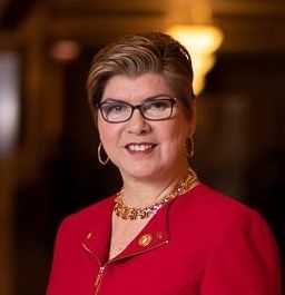 Portrait of a smiling woman in glasses wearing a red jacket and gold necklace against a blurred indoor background