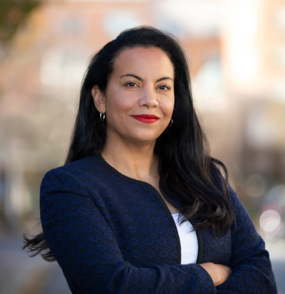 Woman in a navy blazer with folded arms, smiling outdoors against a blurred background.