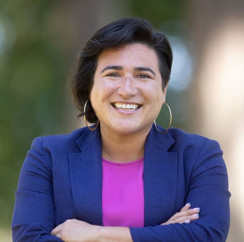 Smiling person in a blue blazer and pink shirt with arms crossed outdoors