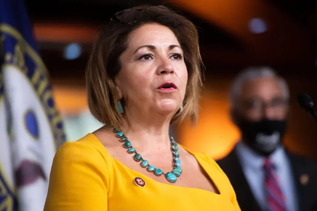 A woman in a yellow dress and turquoise necklace is standing in front of a microphone.