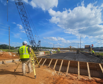 Land Surveying — Meridian, MS — Engineering Plus, Inc. Surveyor in safety vest using a transit on a road construction site with a steamroller in the background.