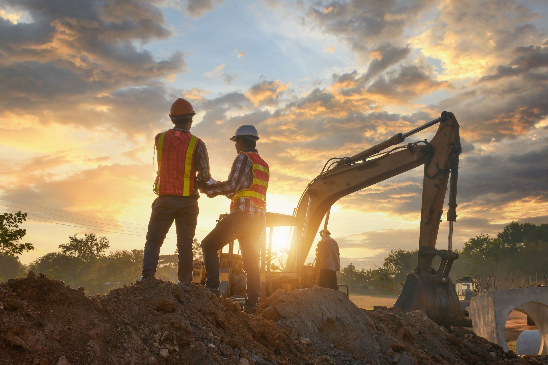 Construction workers in safety vests standing beside an excavator at a sunset job site.