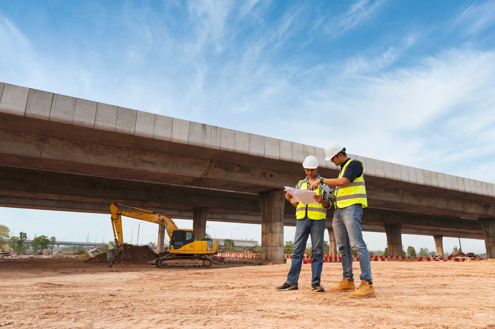 Two construction workers in hard hats stand under a bridge, discussing plans with tools in hand. Two construction workers in hard hats stand under a bridge, discussing plans with tools in hand.
