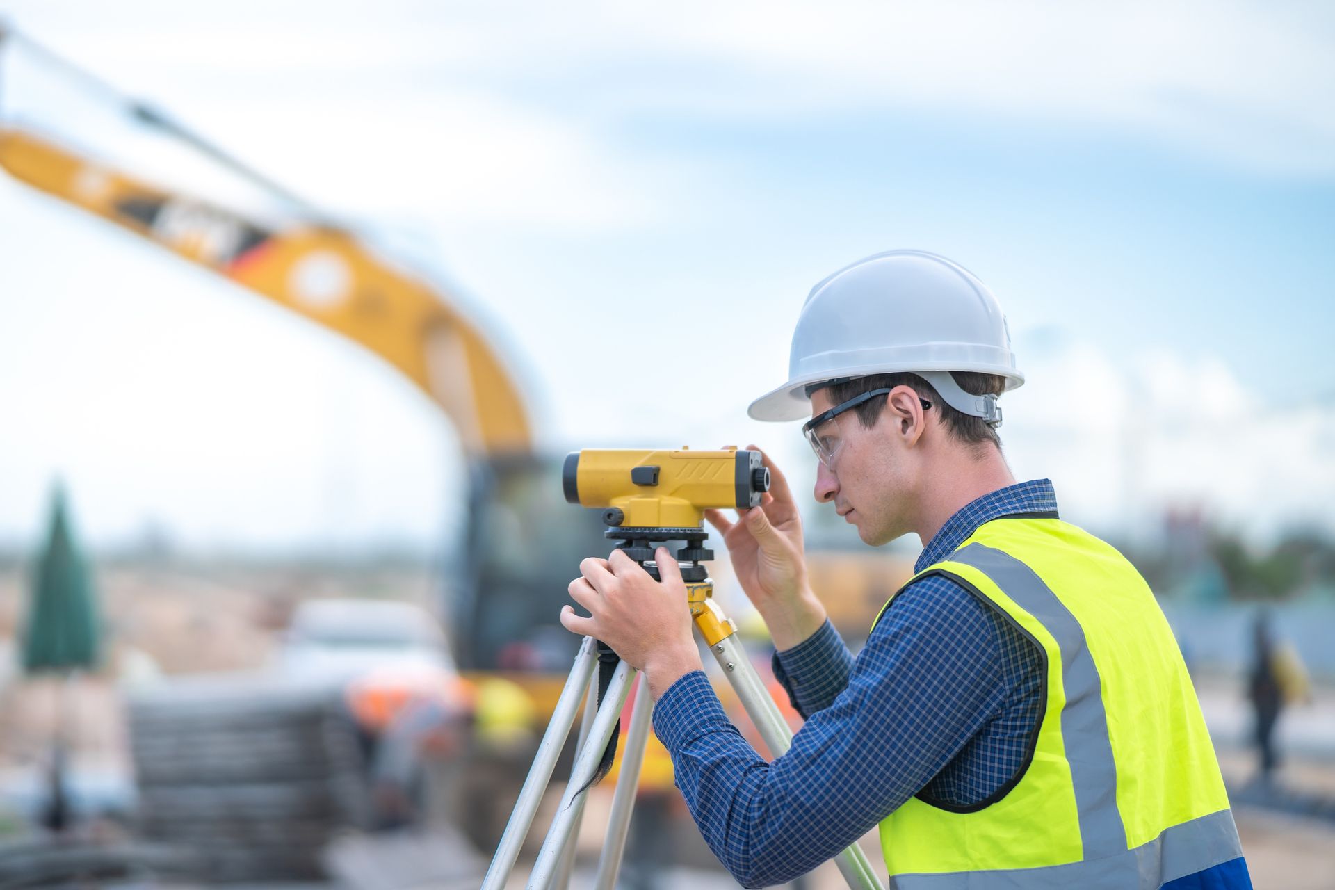 Surveyor in hard hat and safety vest uses a level on a tripod at a construction site.