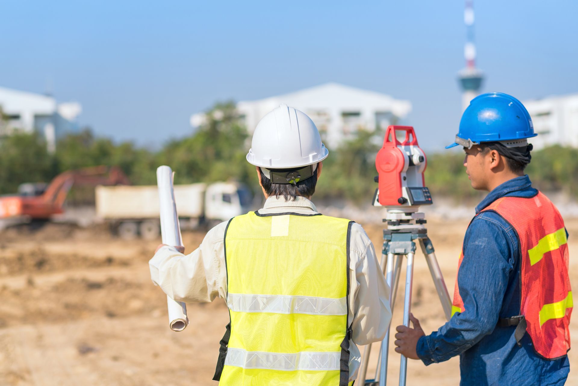 Two construction workers surveying a site with equipment, wearing hard hats and vests.