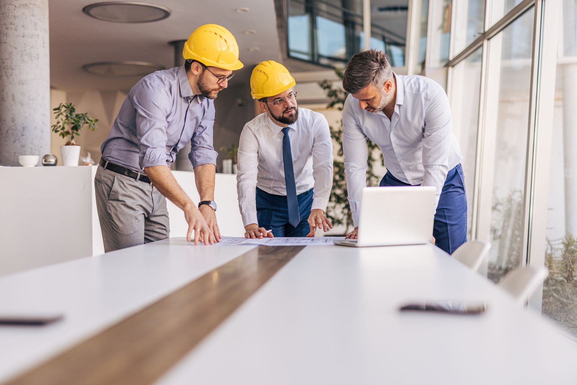 Three men in hard hats review blueprints and a laptop at a conference table.