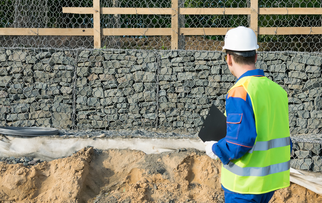 A construction worker in a hard hat and high-visibility vest stands by a gabion wall and a pile of dirt.