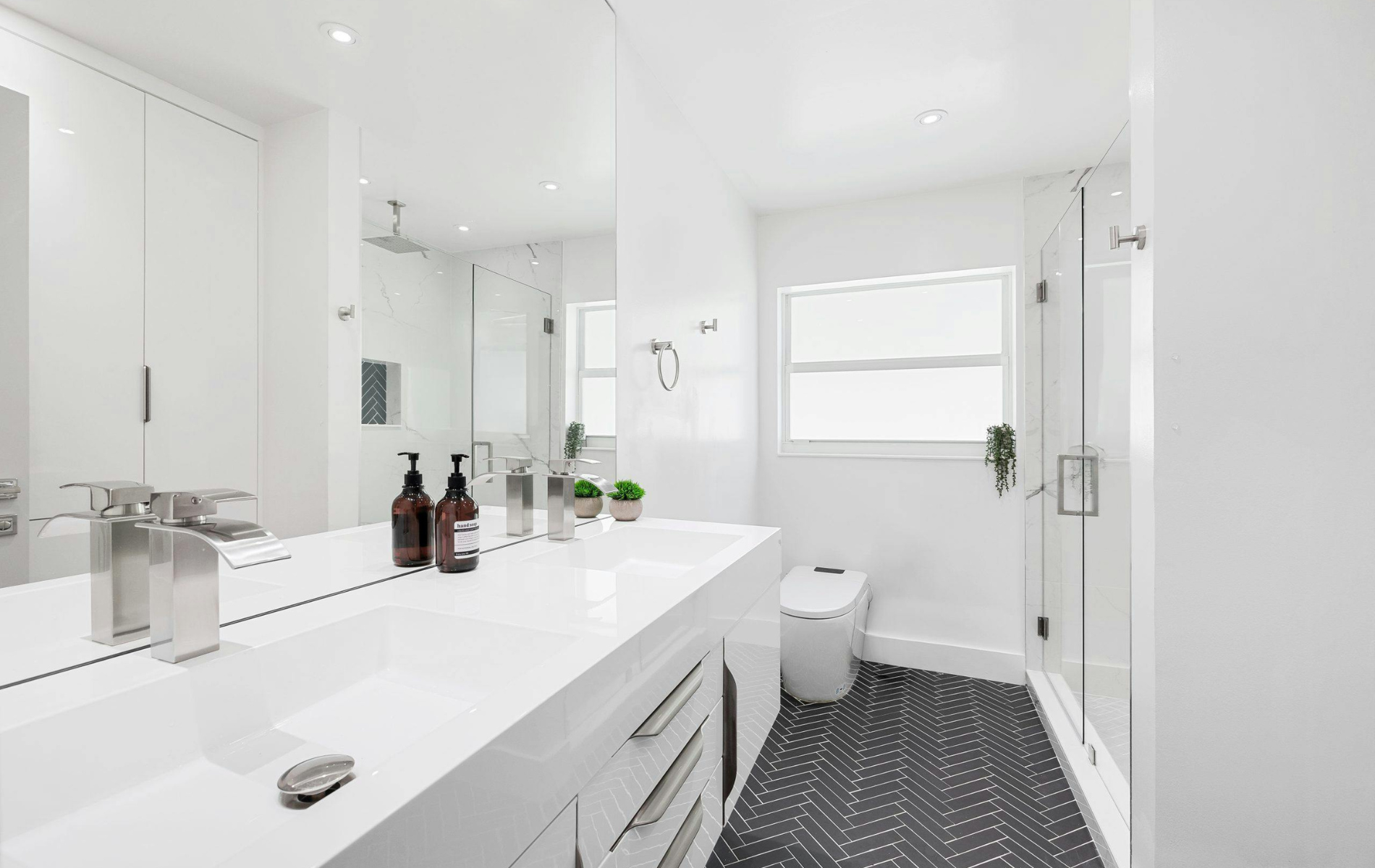 Modern white bathroom with a long double vanity, a large mirror, a glass-enclosed shower, and patterned dark flooring.