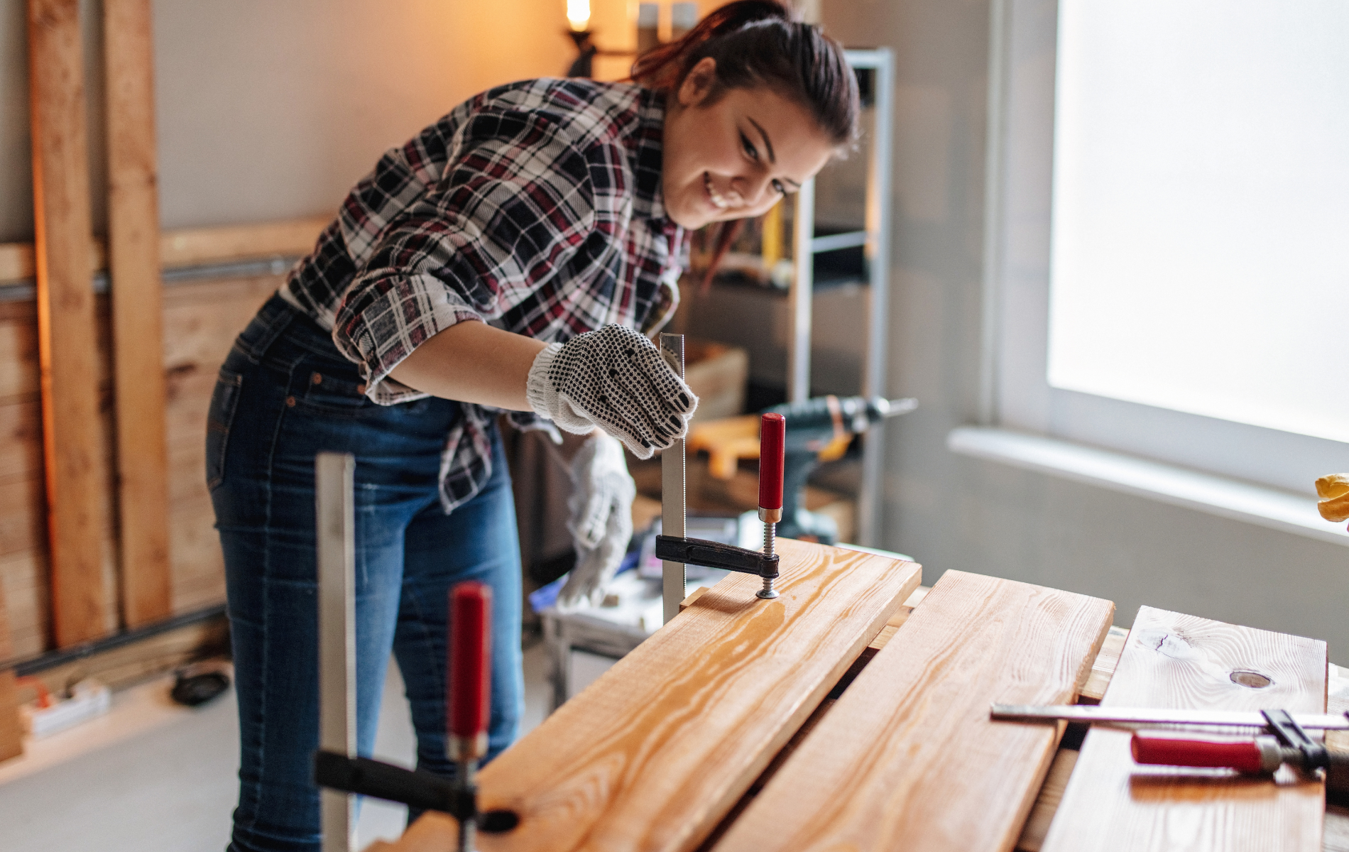 A person wearing plaid and gloves smiles while using a clamp to secure wooden boards on a workbench in a workshop.