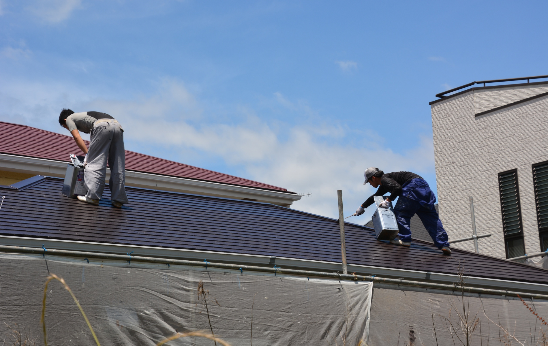 Two workers in work clothes paint a brown metal roof under a bright blue sky.