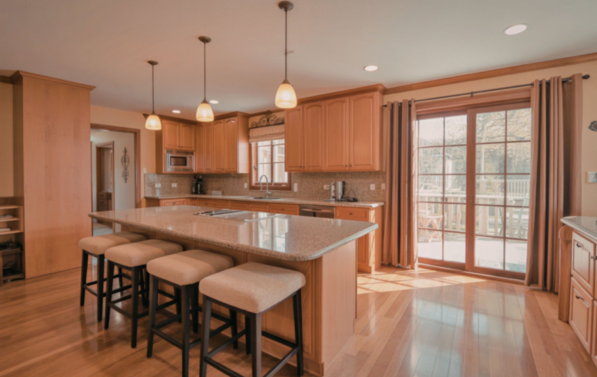 A modern kitchen with a stone-topped island, bar stools, light wood cabinets, and large sliding glass doors.
