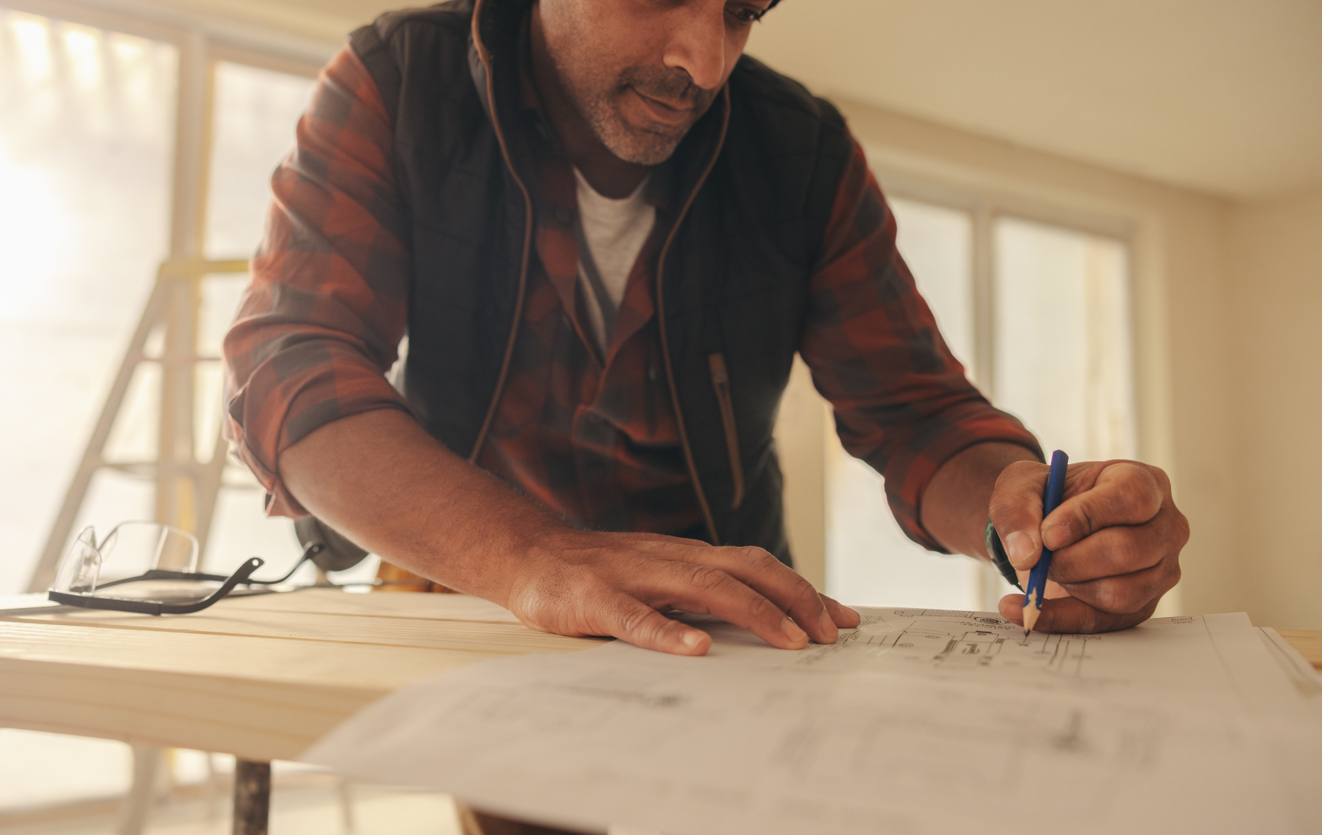 A person in a flannel shirt and vest leans over a wooden surface, using a pencil to mark a blueprint in a sunlit room.
