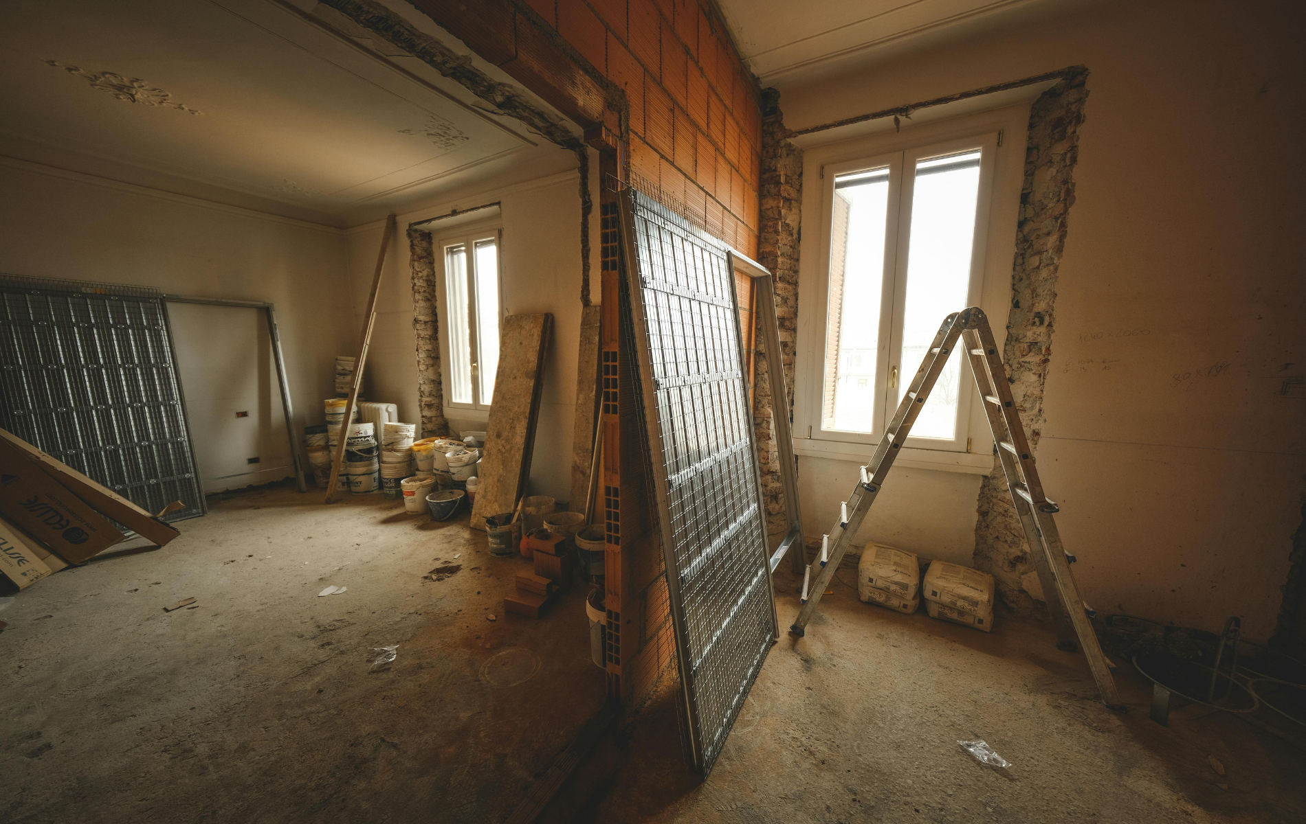 A sunlit room under renovation, featuring exposed brick, scattered building materials, a stepladder, and metal panels.