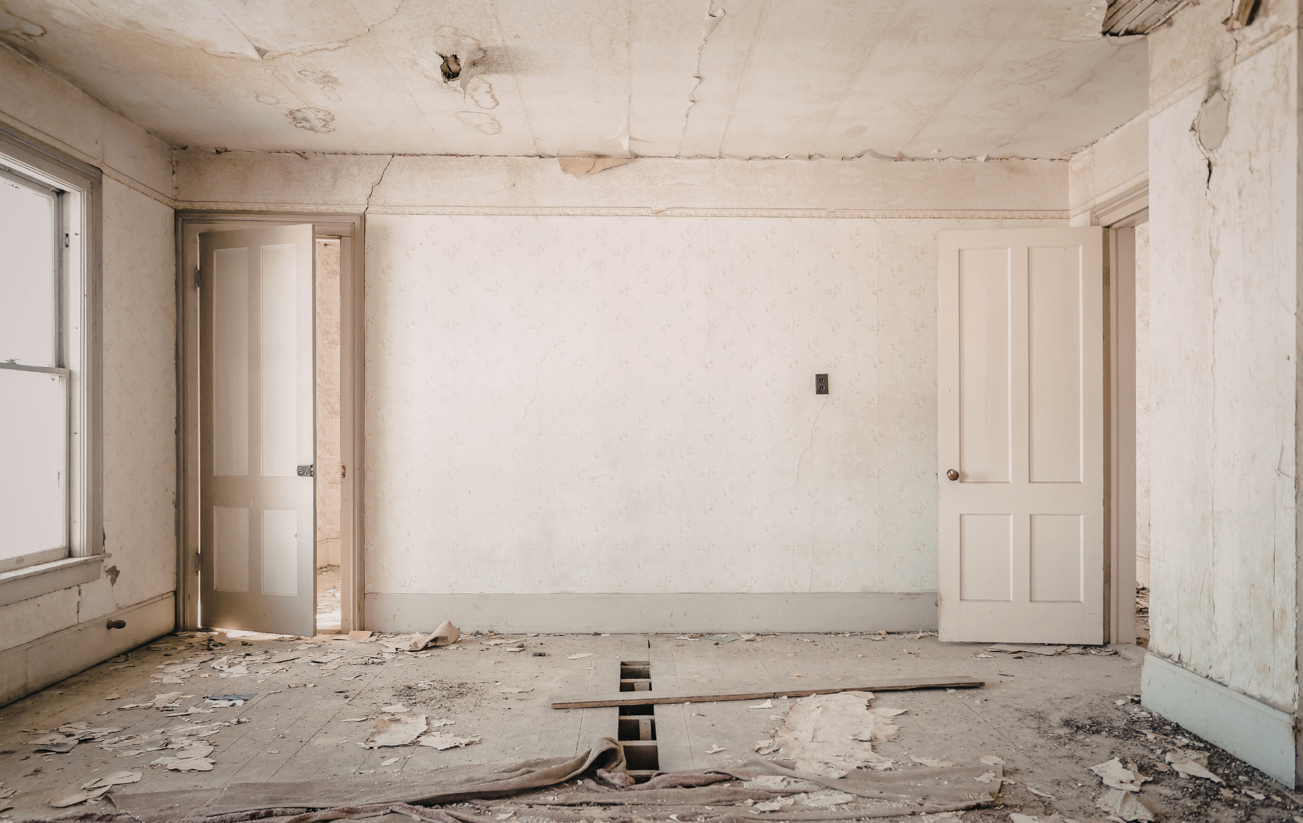 An empty, dilapidated room with peeling white walls, debris-covered floors, and two open doorways.