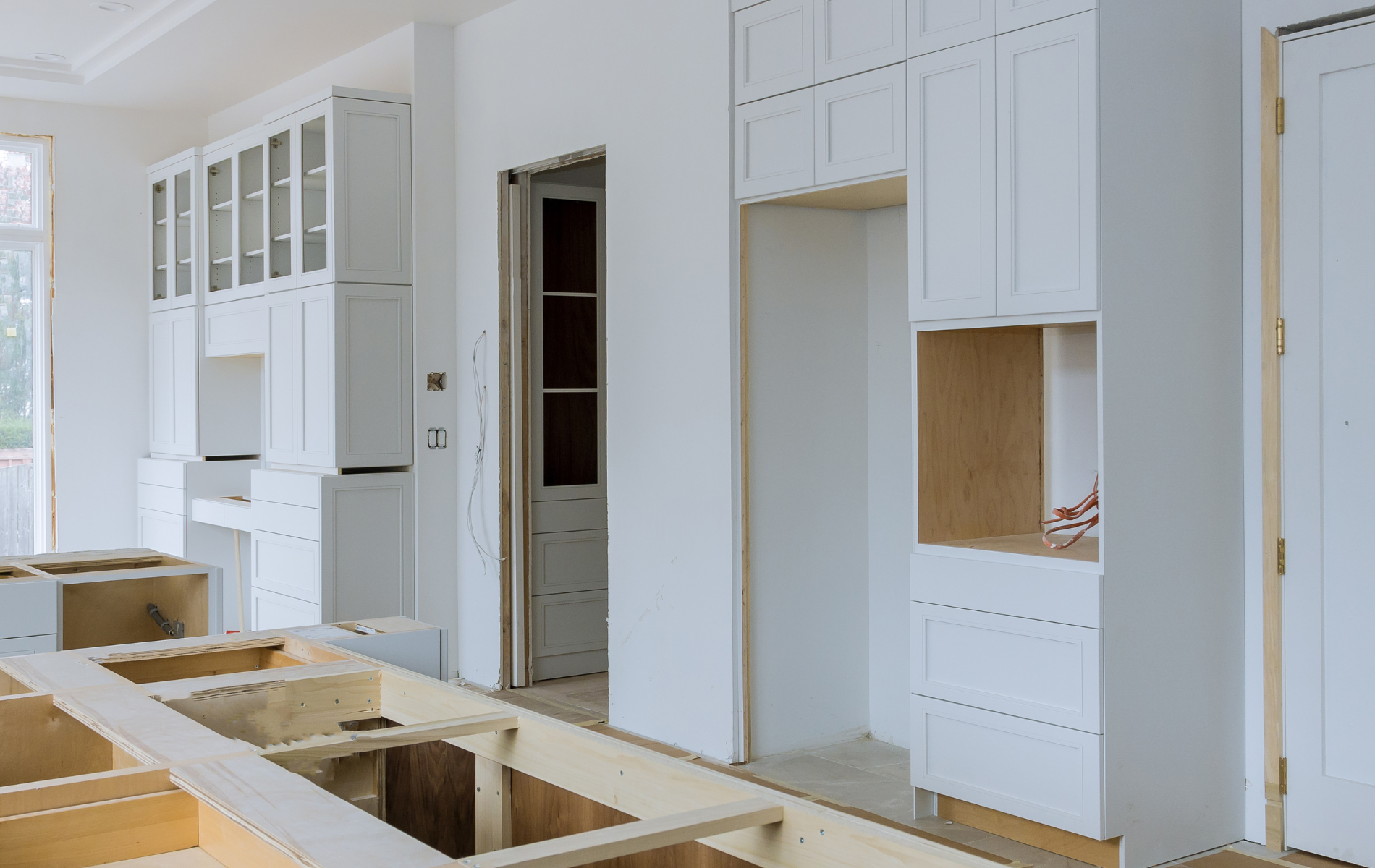 White kitchen cabinets and a wooden island frame under construction in a bright, modern room.