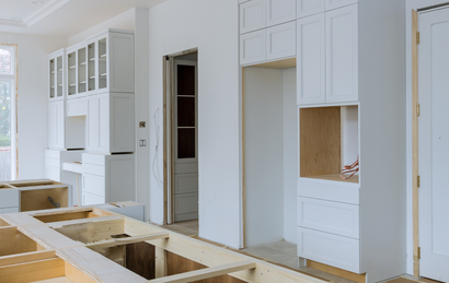 A partially installed kitchen with white cabinets, an island frame, and empty appliance openings during renovation.