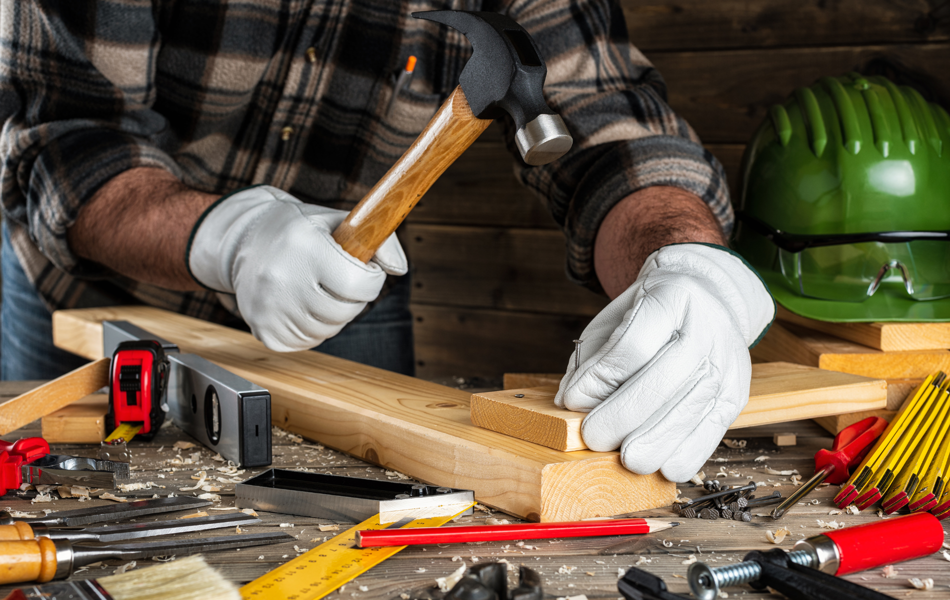 A person wearing work gloves hammers a nail into a wooden board on a workbench covered in tools and equipment.