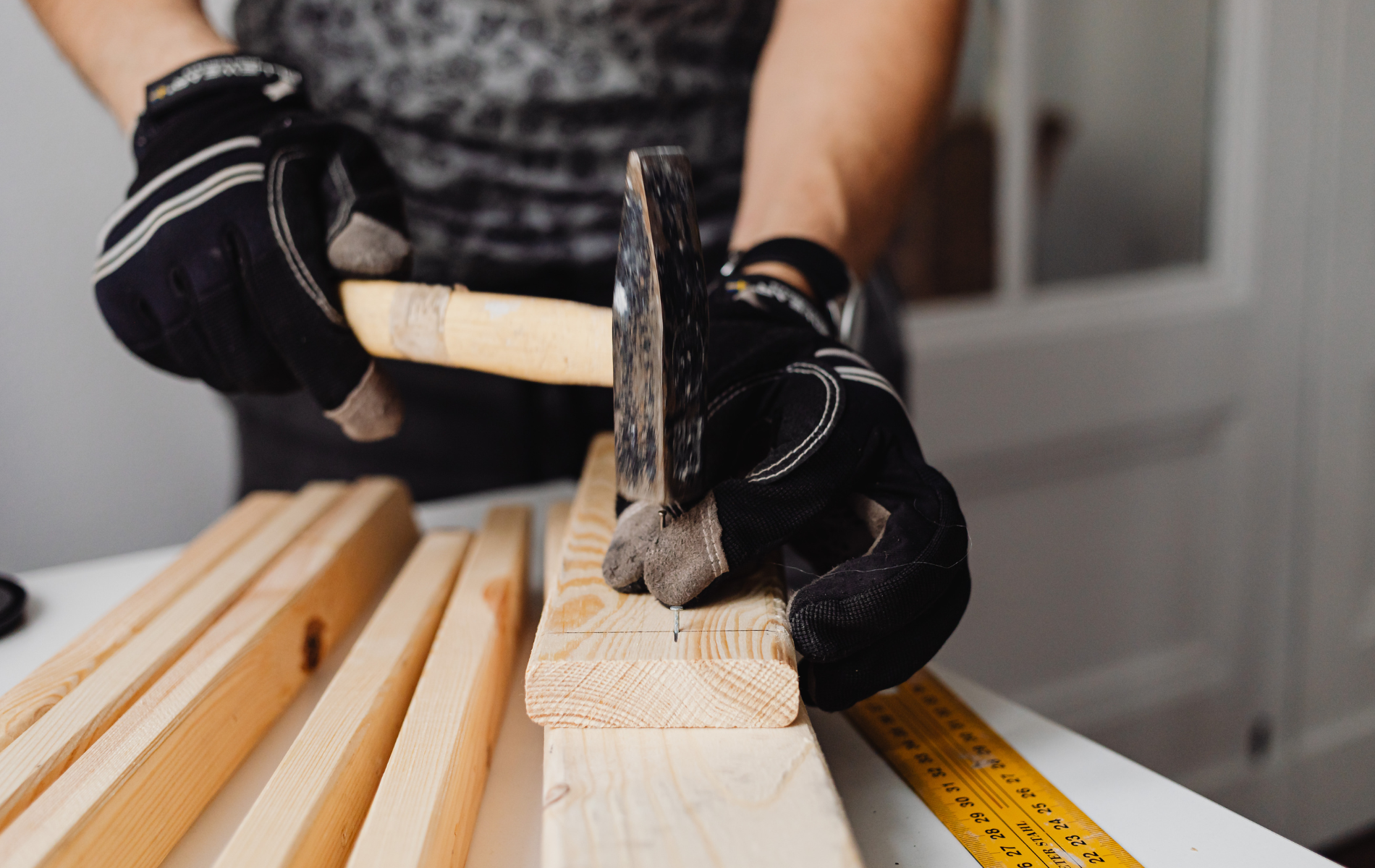 A person wearing black work gloves uses a hammer to drive a nail into a wooden board on a work surface.
