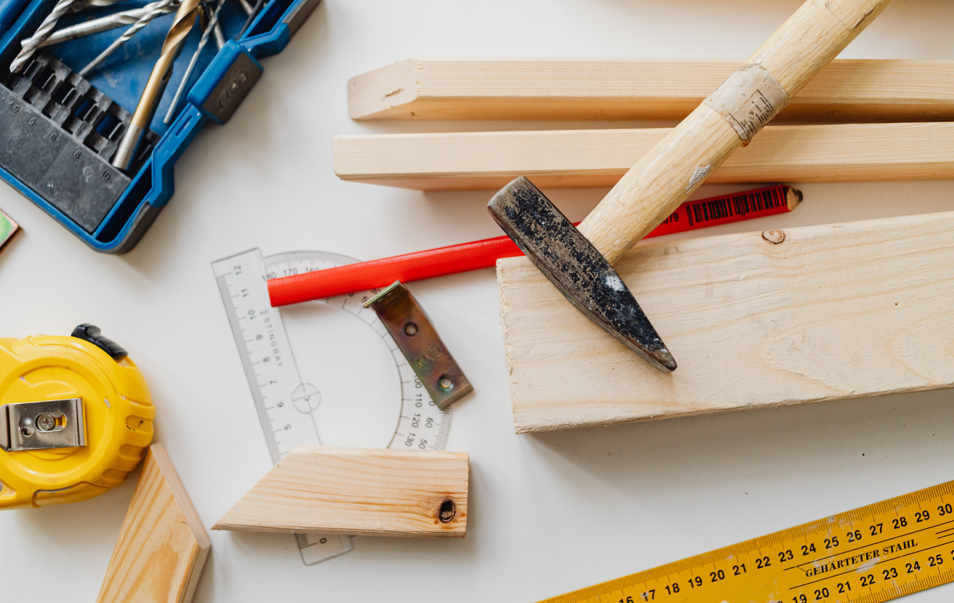 Woodworking tools, including a hammer, tape measure, pencil, and pieces of wood, arranged on a white surface.