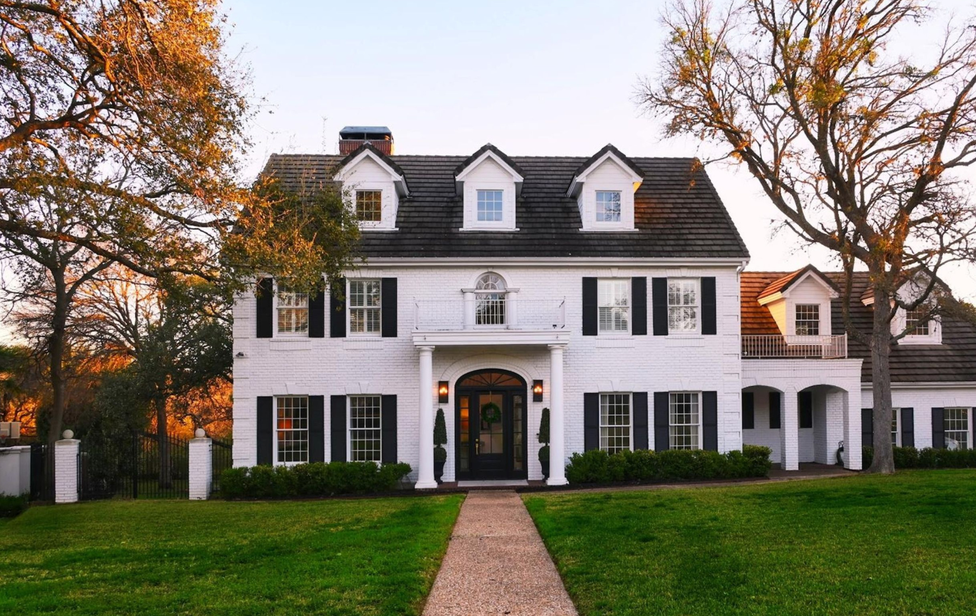 A two-story white colonial-style house with black shutters, dormer windows, and a brick path leading to the front entrance.