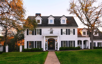A two-story white colonial-style house with black shutters, dormer windows, and a brick path leading to the front entrance.