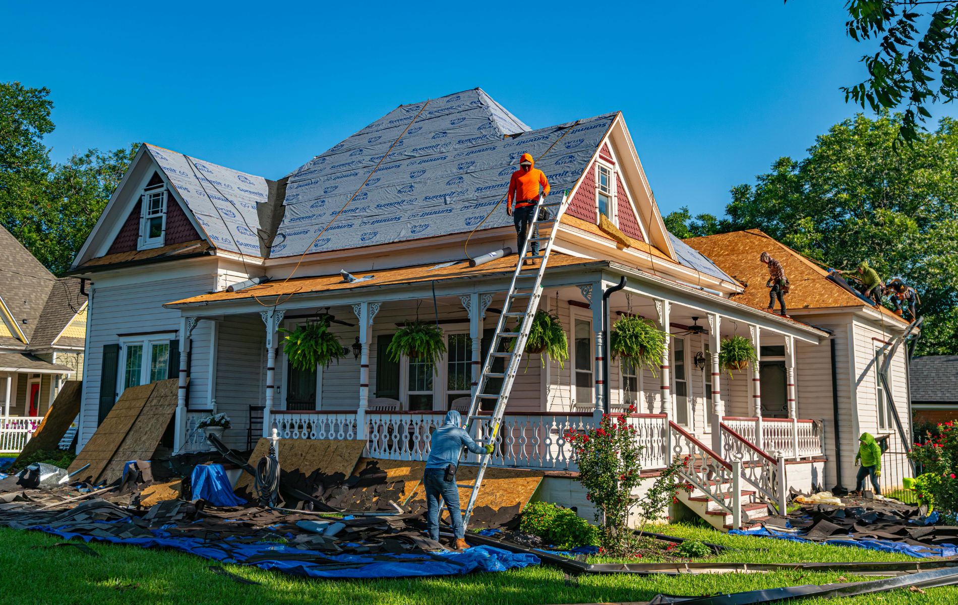 Workers perform roof repairs on a vintage house with a wrap-around porch under a bright blue sky.
