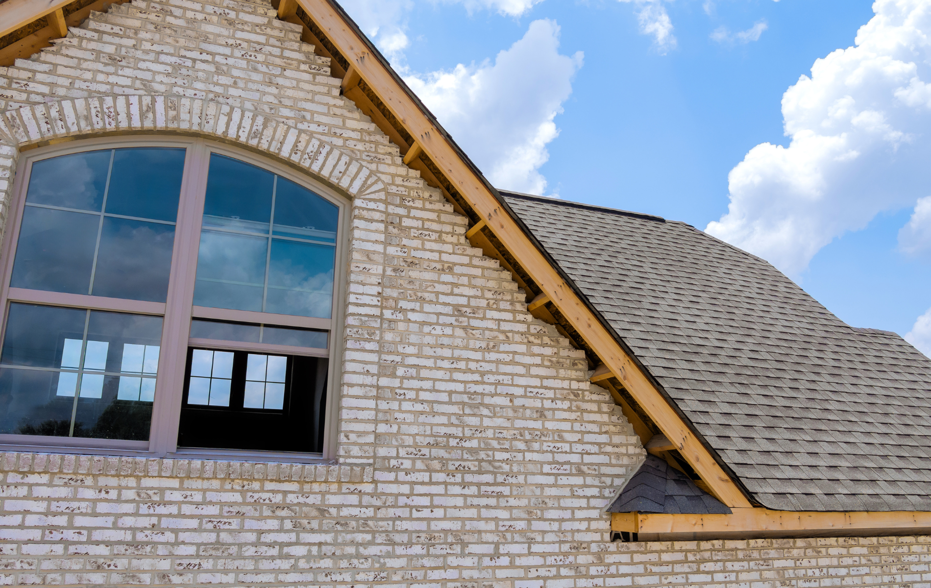 A brick-walled house under a blue sky, featuring a large arched window with paper notes and an unfinished wooden roof eave.