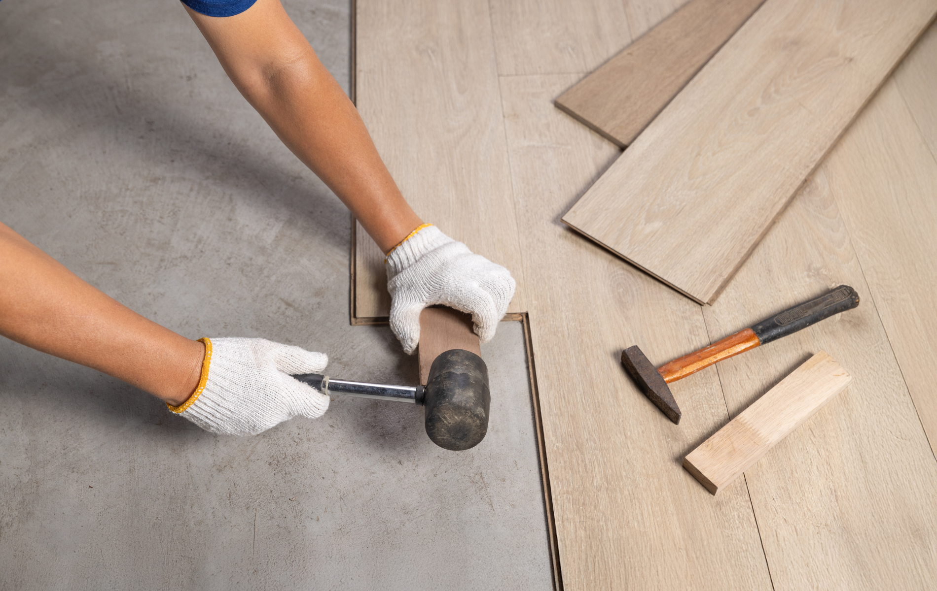 A worker in gloves uses a rubber mallet to install light-colored laminate floor planks on a concrete subfloor.