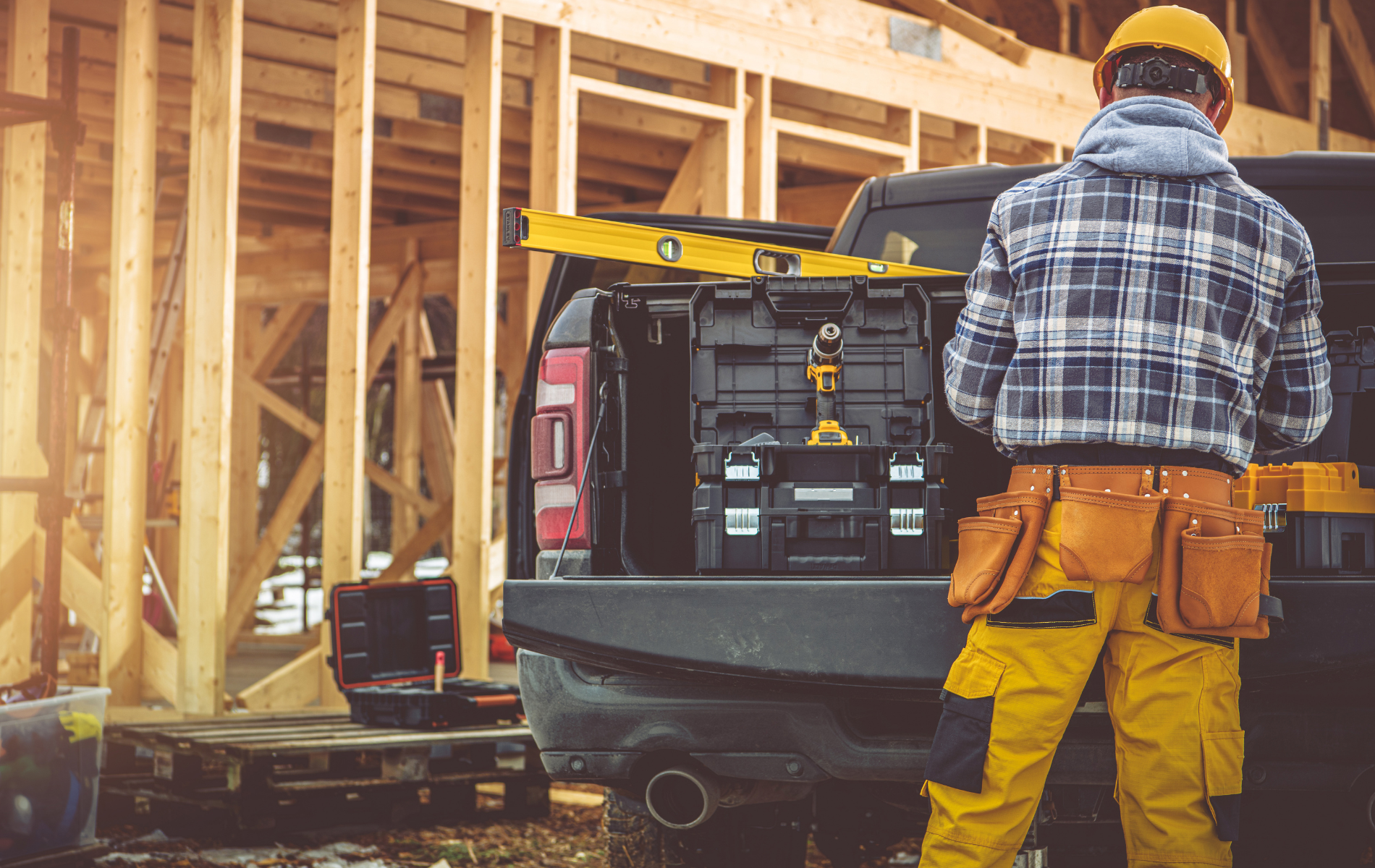 A construction worker in a hard hat and yellow pants stands by a truck bed filled with tool cases at a building site.