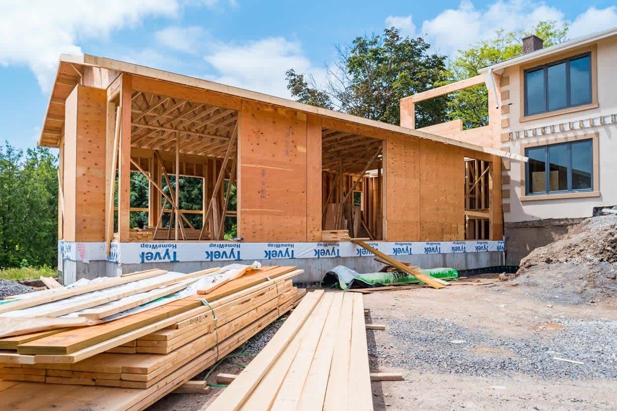 A wooden house addition under construction with stacks of lumber in the foreground against a blue sky.