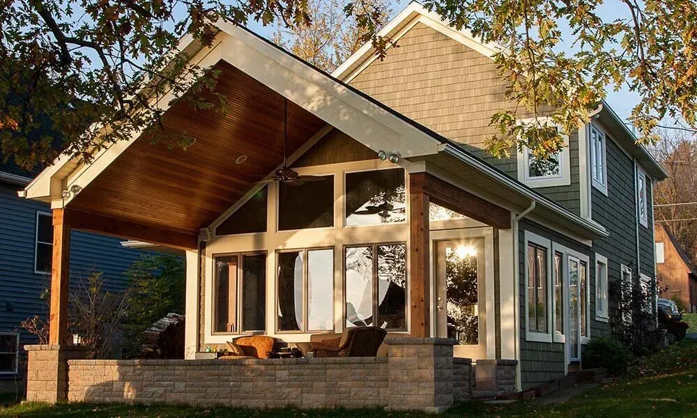 A house with a large, gabled wood-paneled covered patio, large windows, and a stone-walled porch in a residential setting.