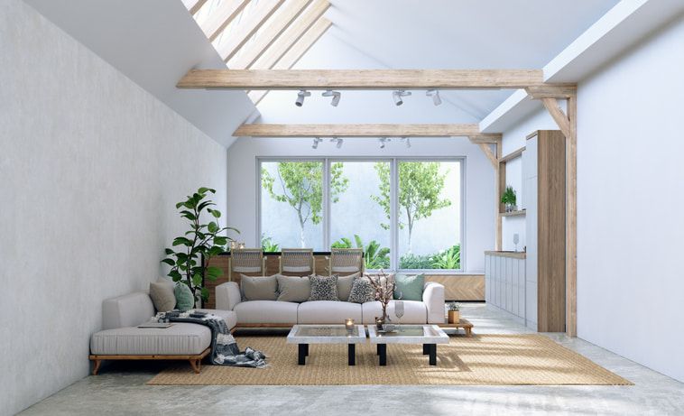 A bright, modern living room with a beige sofa, two coffee tables, a potted plant, and large windows with garden views.