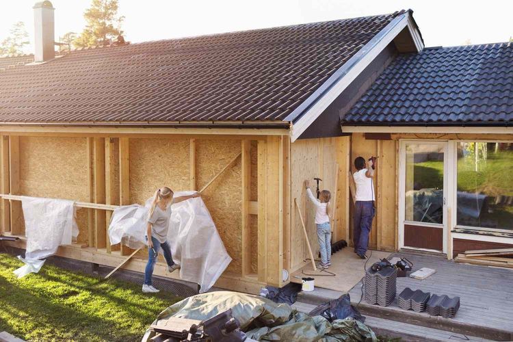 Two people work on a house renovation, fastening wooden panels to an exterior wall frame near a glass door.