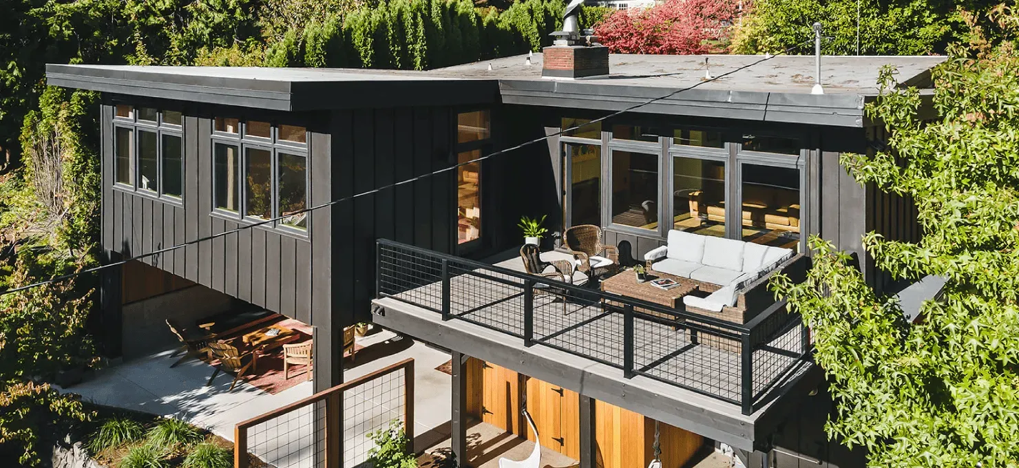 A modern two-story home with dark charcoal siding, a large raised balcony, and a ground-level patio nestled in trees.